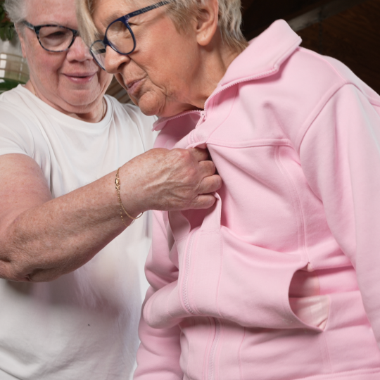 Two elderly women, one helping the other transfer with a pink jacket.